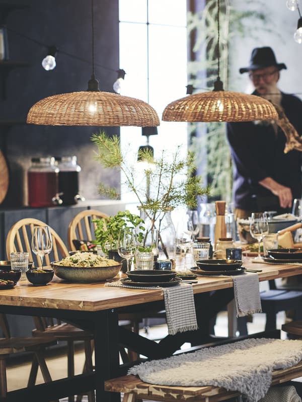 A long table is laid for a meal and is lined with an assortment of chairs, stools and benches of different shapes and sizes. Two pendant lamp shades above.