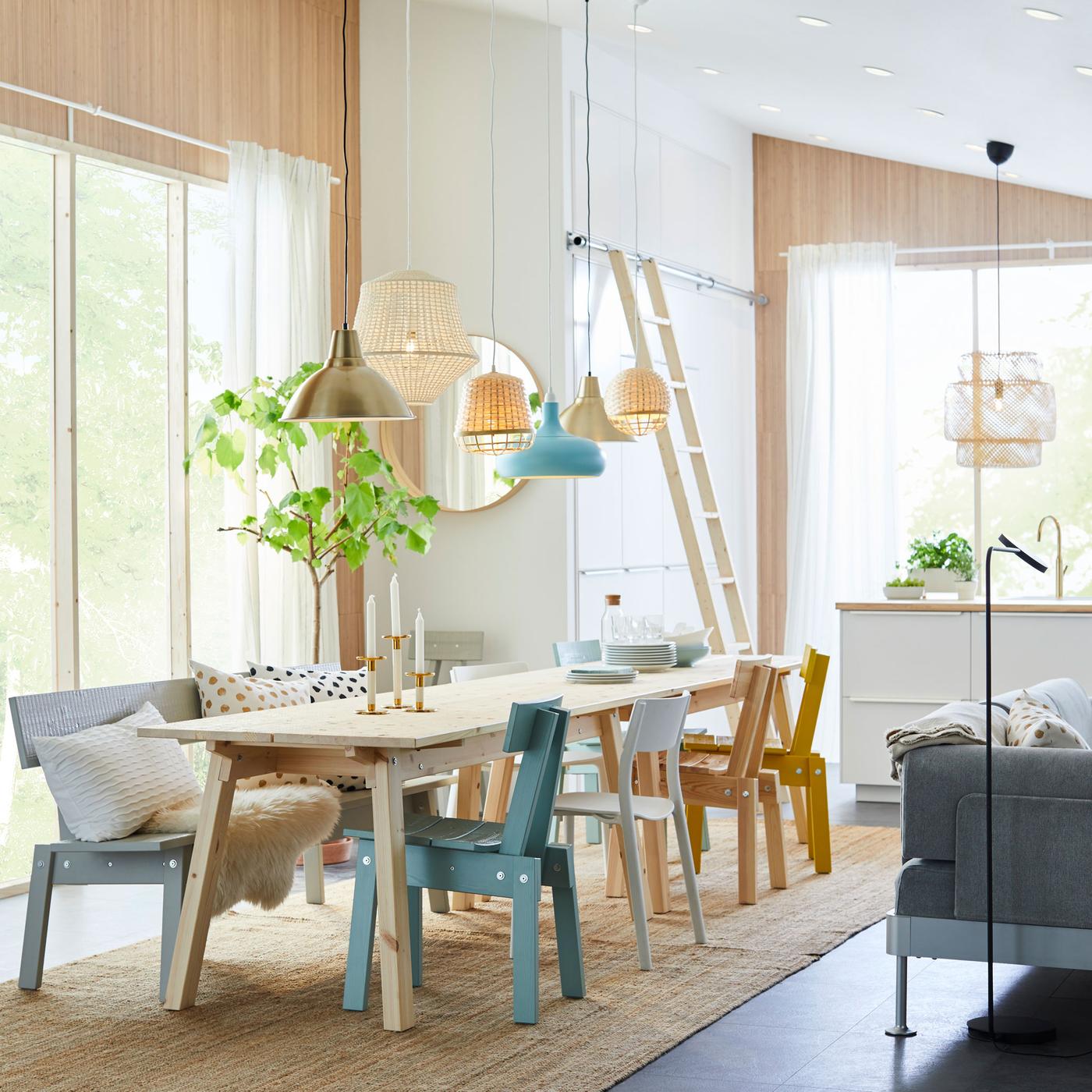 A long wooden dining table with several different color dining chairs next to a white kitchen and a large window.
