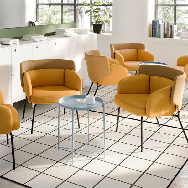 A lounge area with two groups of yellow armchairs, round small metal tables in the middle and a window behind them.