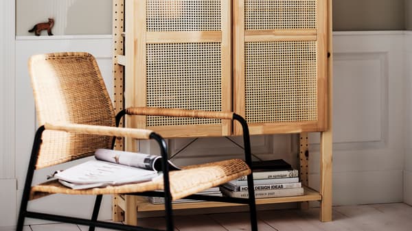 A magazine lying on a rattan/anthracite ULRIKSBERG armchair in front of a pine IVAR shelving unit with doors.