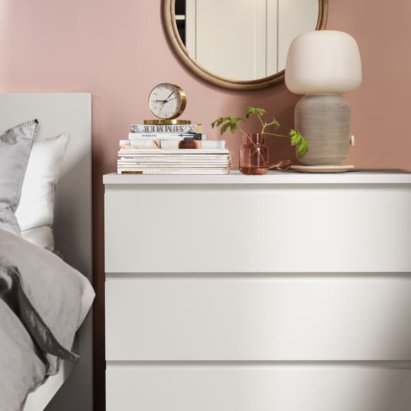 A MALM chest of drawers next to a bed with books, a clock, a lamp and a plant on top of it with a mirror hanging on the wall adjacent to it