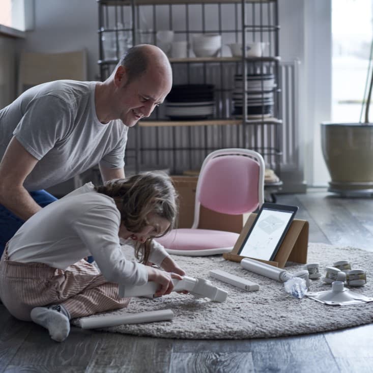 A man and a child, smiling and focused, are assembling white pipes on a circular mat. A tablet and various tools are nearby, suggesting a collaborative project.