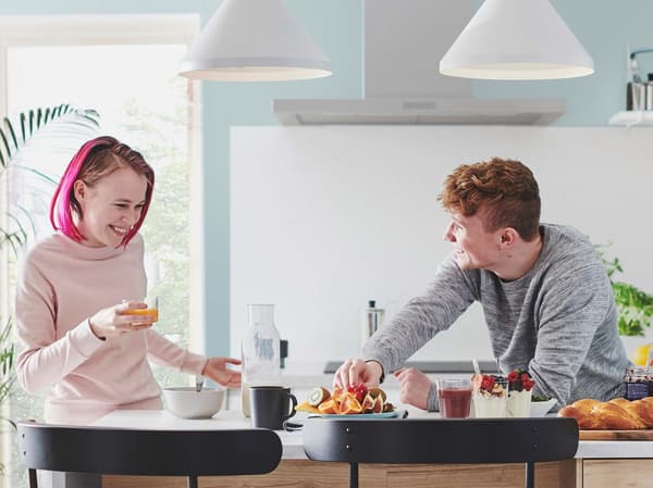A man and a woman are standing behind a kitchen island, tasting the freshly cooked food together and laughing