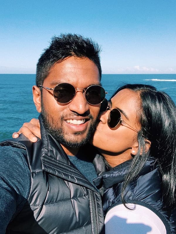 A man and a woman, both wearing sunglasses, are standing in front of the ocean. The woman is kissing the man on his cheek.