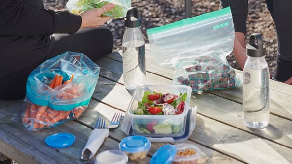 A man and a woman by a picnic table in a forest, unpacking food placed in ISTAD resealable bags, and IKEA 365+ bottles.