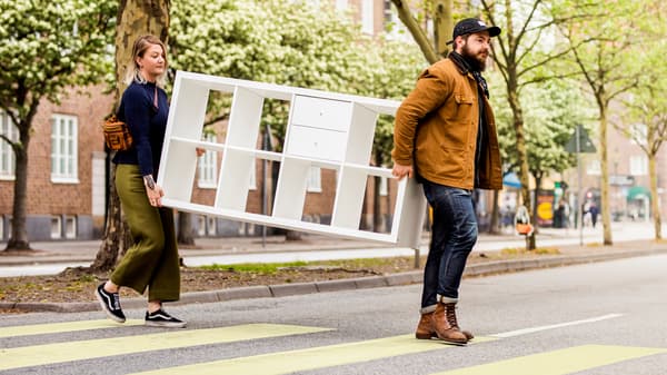 A man and a woman carry a white KALLAX storage unit across a street.