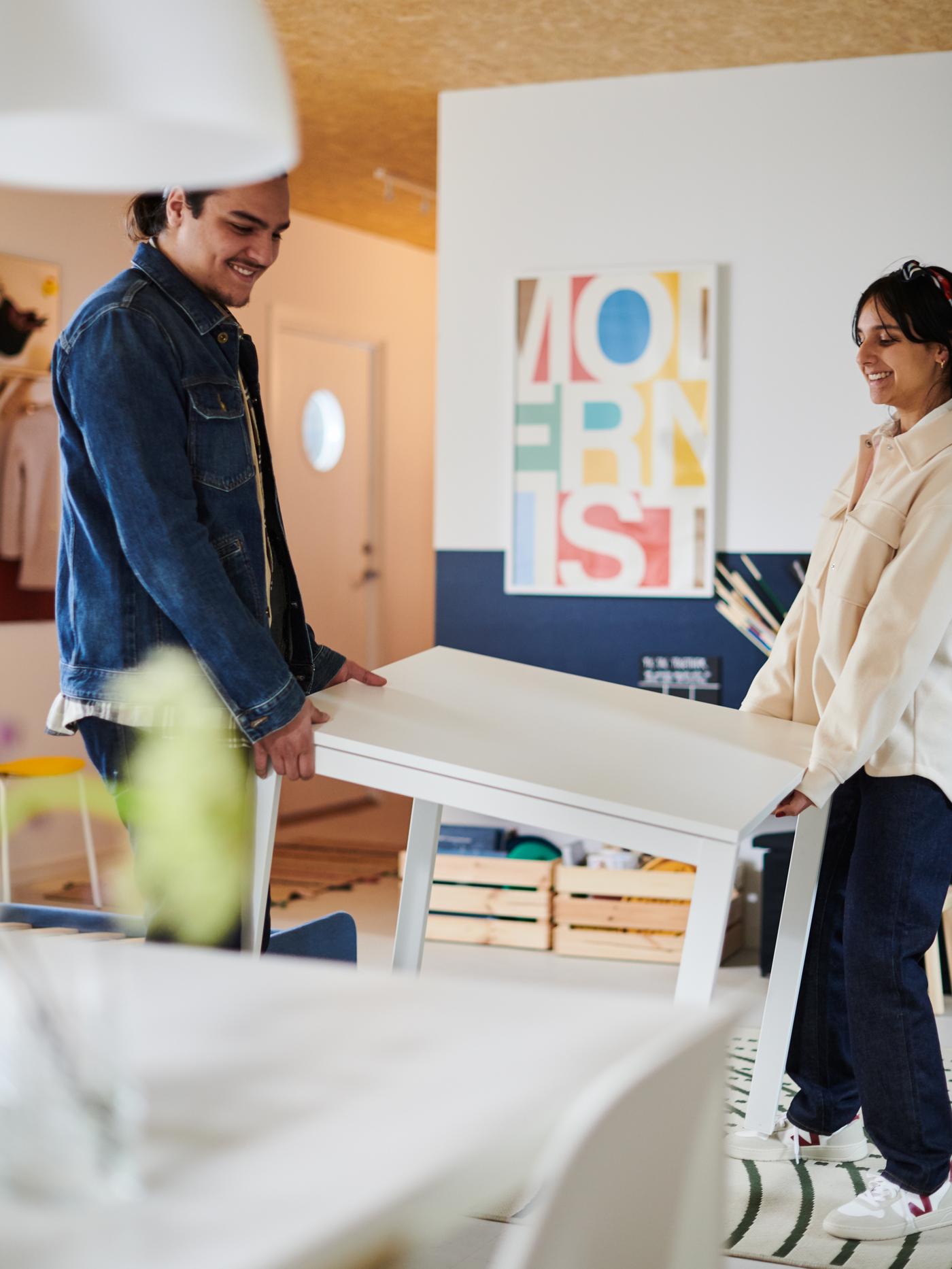 A man and a woman moving a white MELLTORP table, with two pine KNAGGLIG boxes behind and a white MELODI pendant above.