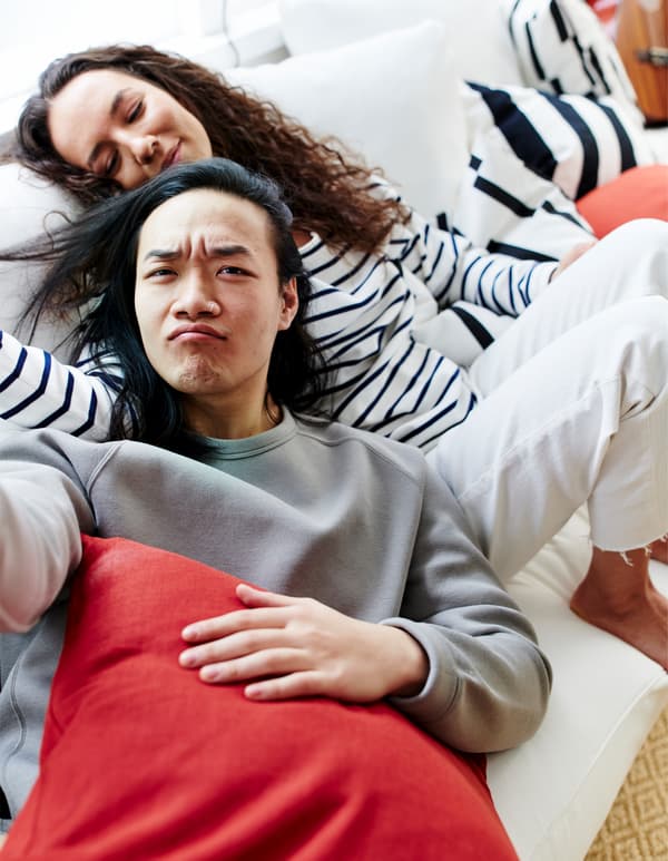 A man and a woman nestled together on a white sofa, surrounded by black-and-white striped and coloured cushions.