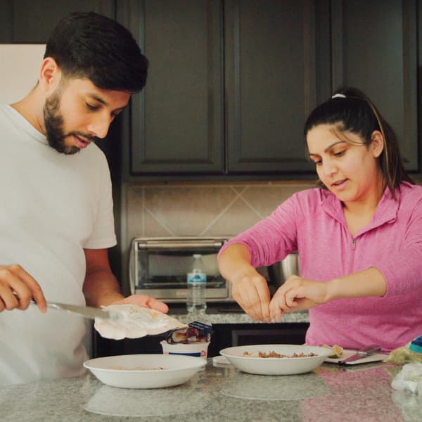 A man and a woman with one plate each are preparing breakfast in a kitchen with a stone worktop and grey cabinets.