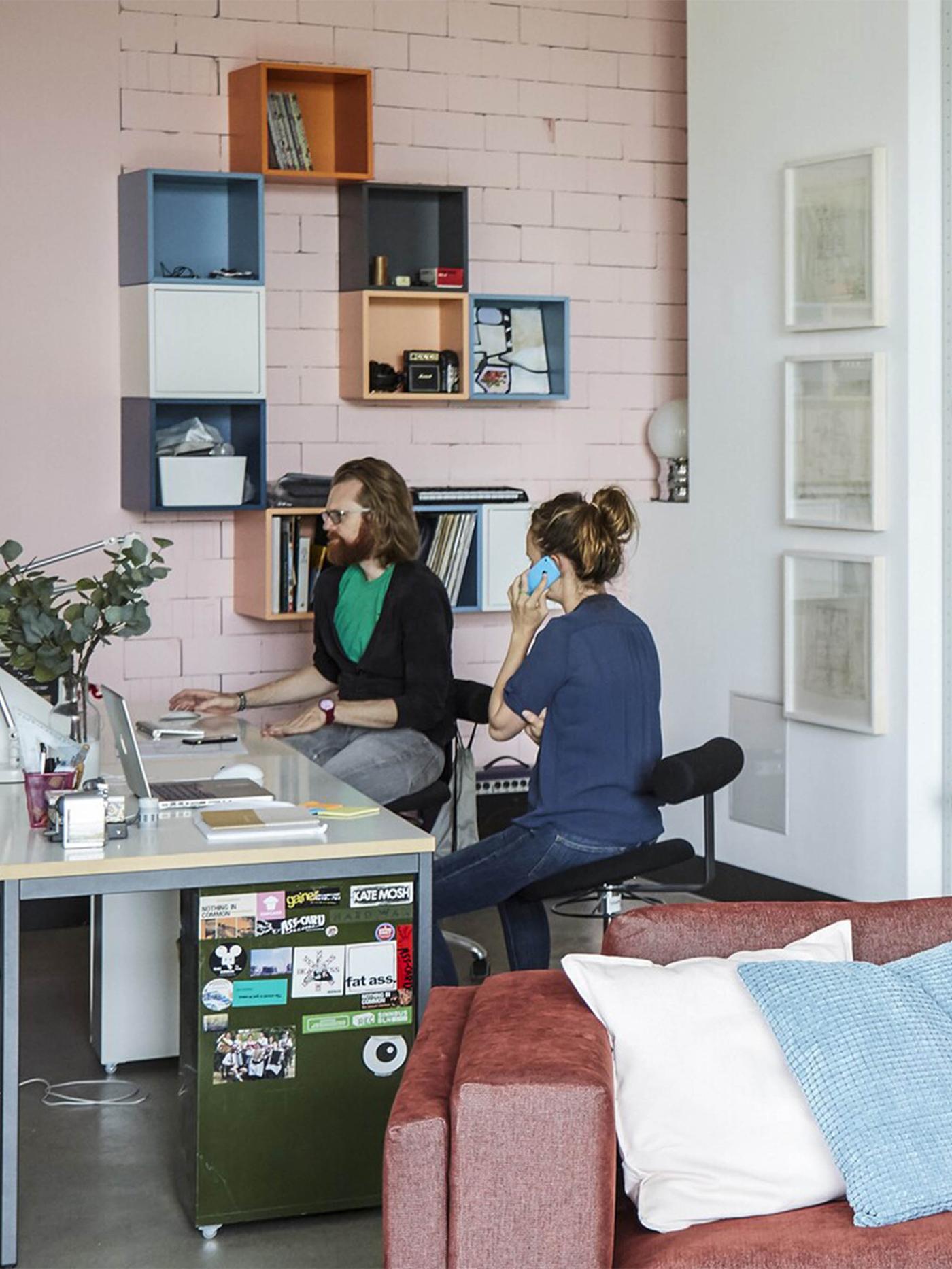 A man and a woman working at their desks in the corner of the big, open space living room with light pink brick walls.