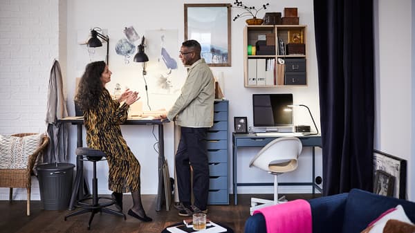 A man and woman in a home office. He stands next to an IDÅSEN sit/stand desk, she sits on a LIDKULLEN sit/stand support.