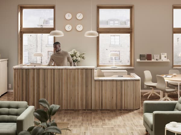 A man at a light wood reception desk in front of two windows. Two NYMÅNE pendants are above and four clocks are on the wall.