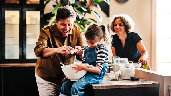 A man cracks an egg into a bowl held by a child sitting on a kitchen worktop while a woman watches them.