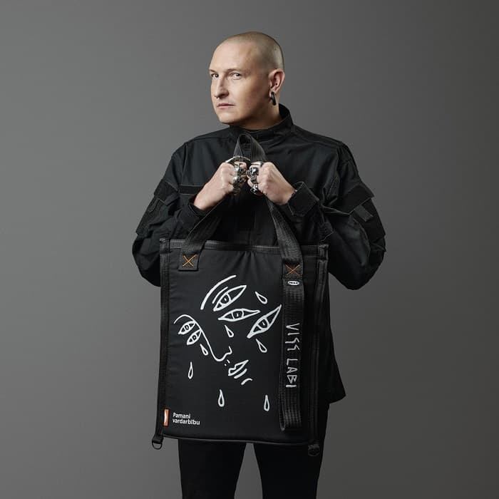 A man dressed in black stands against a dark background, holding a black bag with a graphic image of a face and tears.