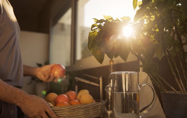 A man fetching taking fresh fruit out of a woven basket to wash them in the kitchen sink, with the sun peeks through a plant.