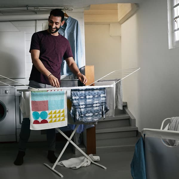 A man hanging t-shirts and towels in a basement room, on an IKEA MULIG drying rack.