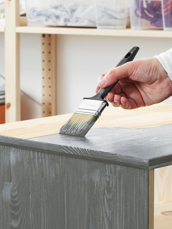 A man holding a paintbrush, painting a piece of furniture gray