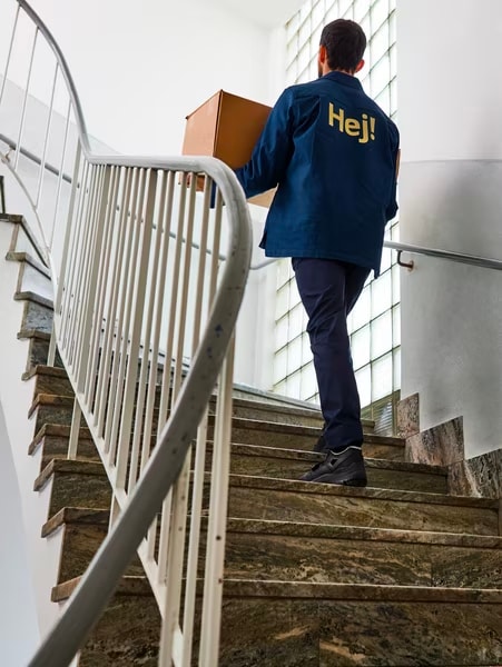 A man in a blue IKEA jacket with the word "Hej!" carries a carton box up a flight of stairs.