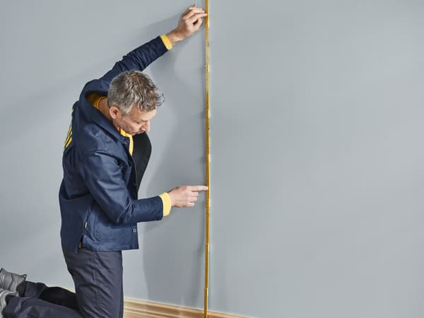 A man in a blue jacket with a measuring tape and measuring along a wall.