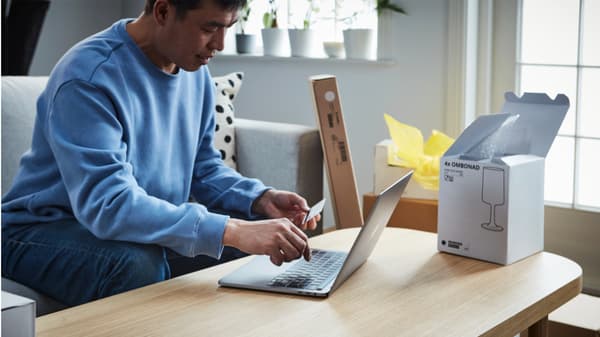 A man in a blue sweater sits at a wooden table, using a laptop and holding a card. Open boxes suggest unpacking. The room is bright and modern.