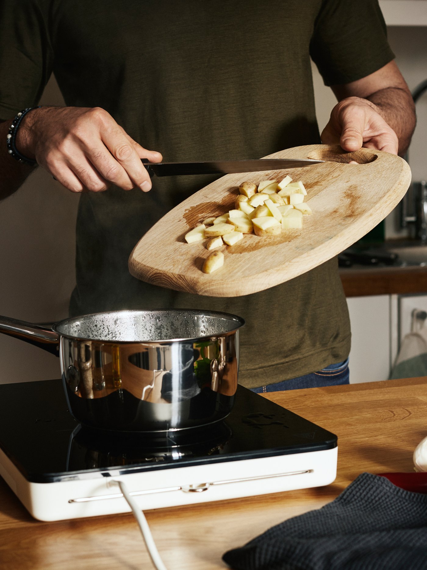 A man in a green t-shirt scrapes diced potato from a wooden chopping board into a pan on a TILLREDA portable induction hob.