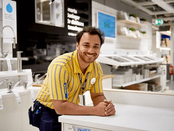 A man in a yellow IKEA uniform leaning on a countertop in an IKEA kitchen department smiling at the camera.
