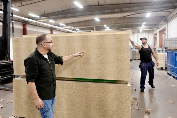 A man in jeans and a dark green shirt stands in a factory room. He stands in front of a stack of particle boards and points to the boards.