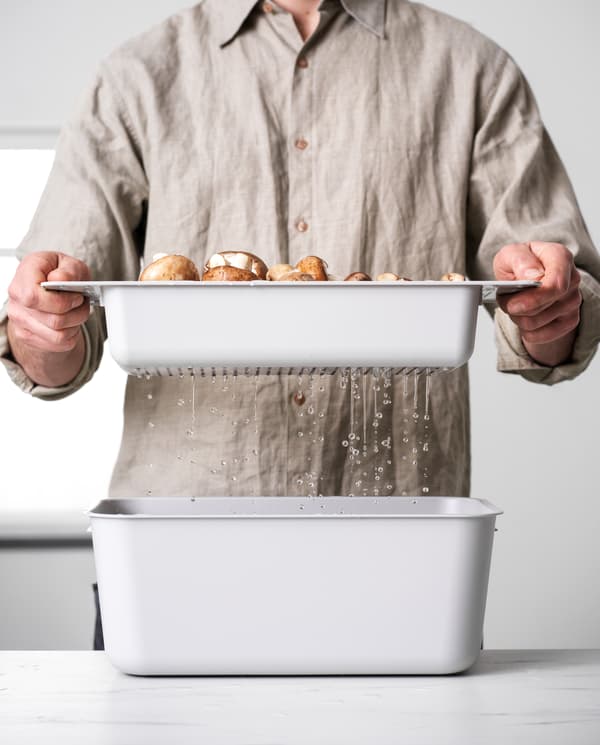 A man is holding a grey colander with mushrooms over a grey washing-up bowl. Water drips down into the washing-up bowl.