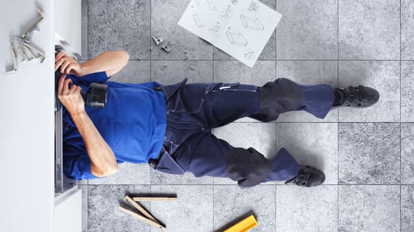 A man is lying on fabric with his head in a cupboard, assembling; on the floor are assembly instructions and tools.