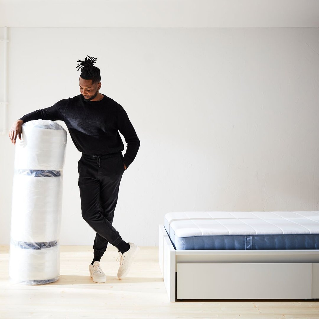 A man is standing next to a rolled-up mattress and a bed with a new mattress on it. 
