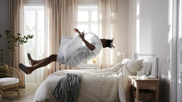 A man jumping into a bed placed in a neutral-styled bedroom with a birch nightstand and the curtains partially open.