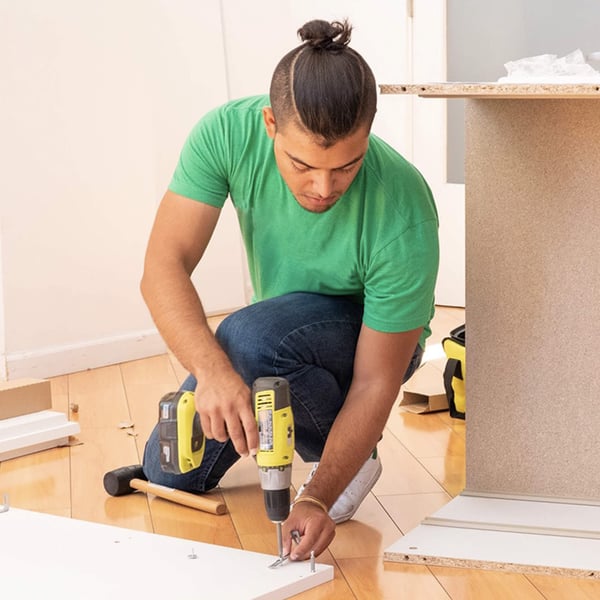 A man kneeling down with a drill, assembling a piece of furniture.