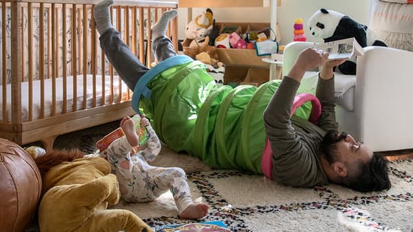 A man laying on the ground with a BUSA play tunnel around his waist and soft toys surrounding him.