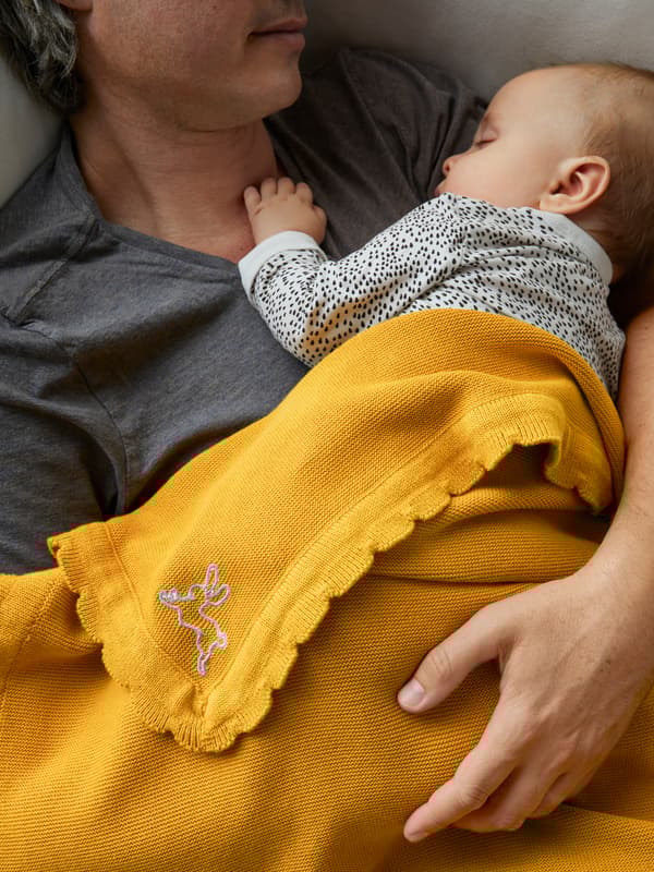 A man lying comfortably in a sofa while holding a sleeping child partly covered by a dark yellow SOLGUL blanket.