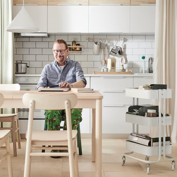 A man opens his computer on a RÖNNINGE extendable table in a kitchen with white VEDDINGE and ash effect ASKERSUND doors.