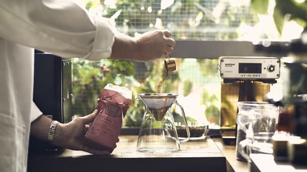 A man scoops coffee grounds into a filter inside a glass coffee pot next to a coffee machine on a kitchen counter top.