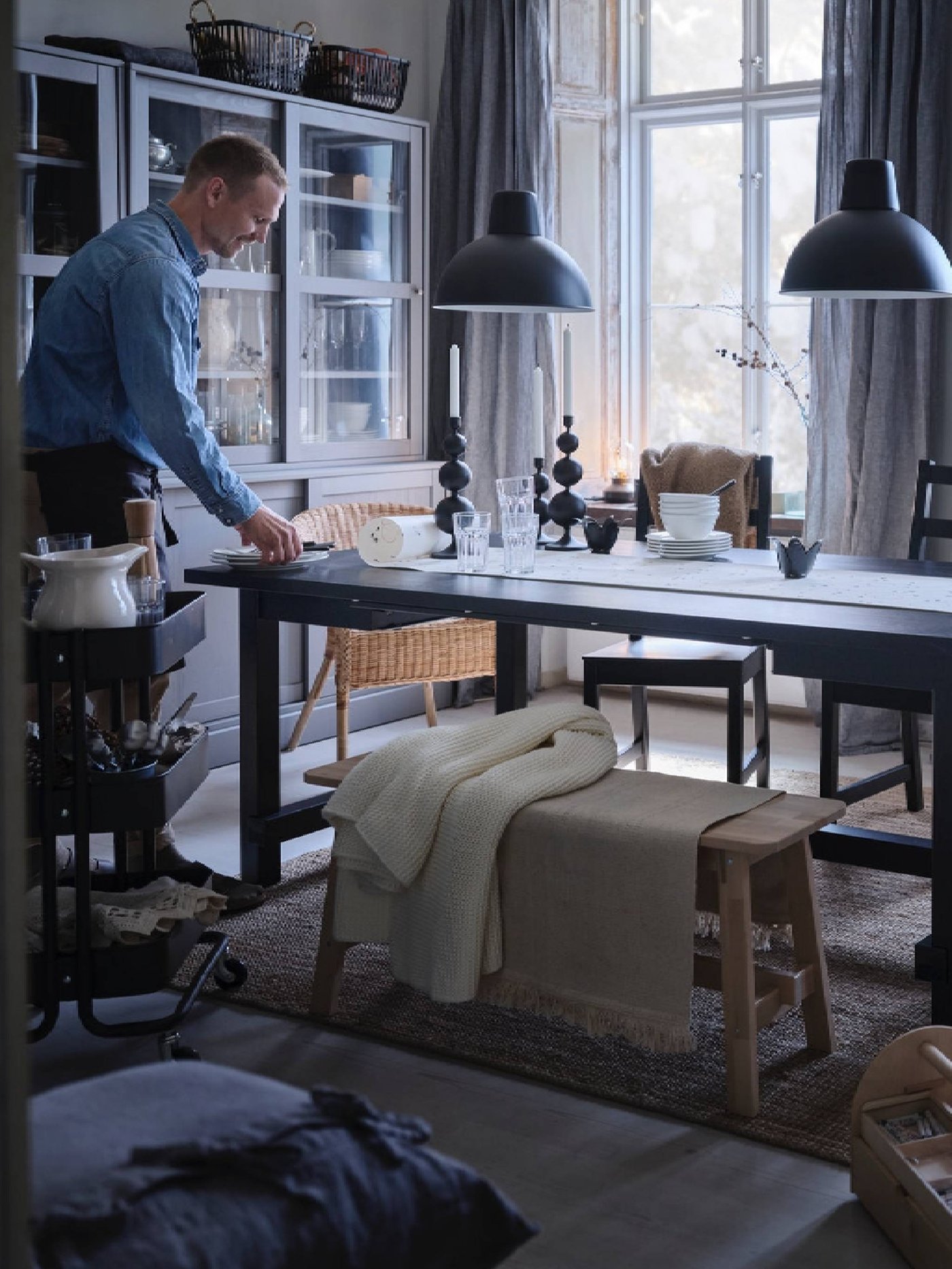 A man sets one end of a table for holiday dinner. Blankets draped over a wooden bench beside the table.