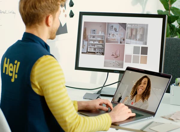 A man sits at a desk engaged in a video call with a woman via a laptop. A second screen shows images of room interiors.