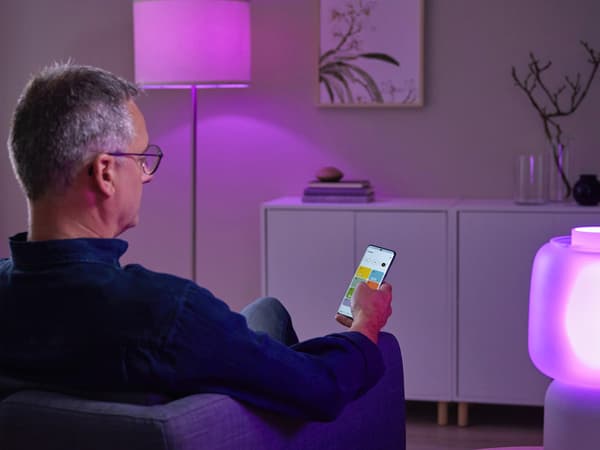 A man sitting in a chair, holding a mobile device, looking at the IKEA Home smart app enabled by DIRIGERA hub.
