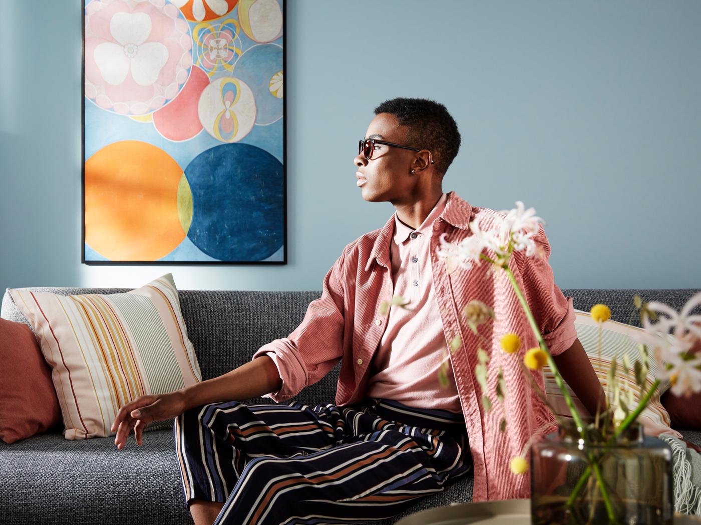 A man sitting in a grey sofa in front of a light-blue wall with a large poster of abstract art in pastel colours.