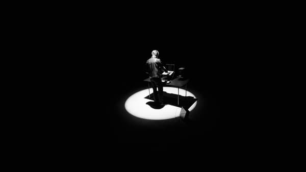 A man standing in black space under a spotlight at the black OBEGRÄNSAD desk with a laptop and speakers.