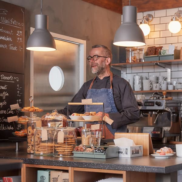 A man standing in the café kitchen, serving trays with fresh bakery, grey ceiling lamps, white coffee mug and wall shelves.