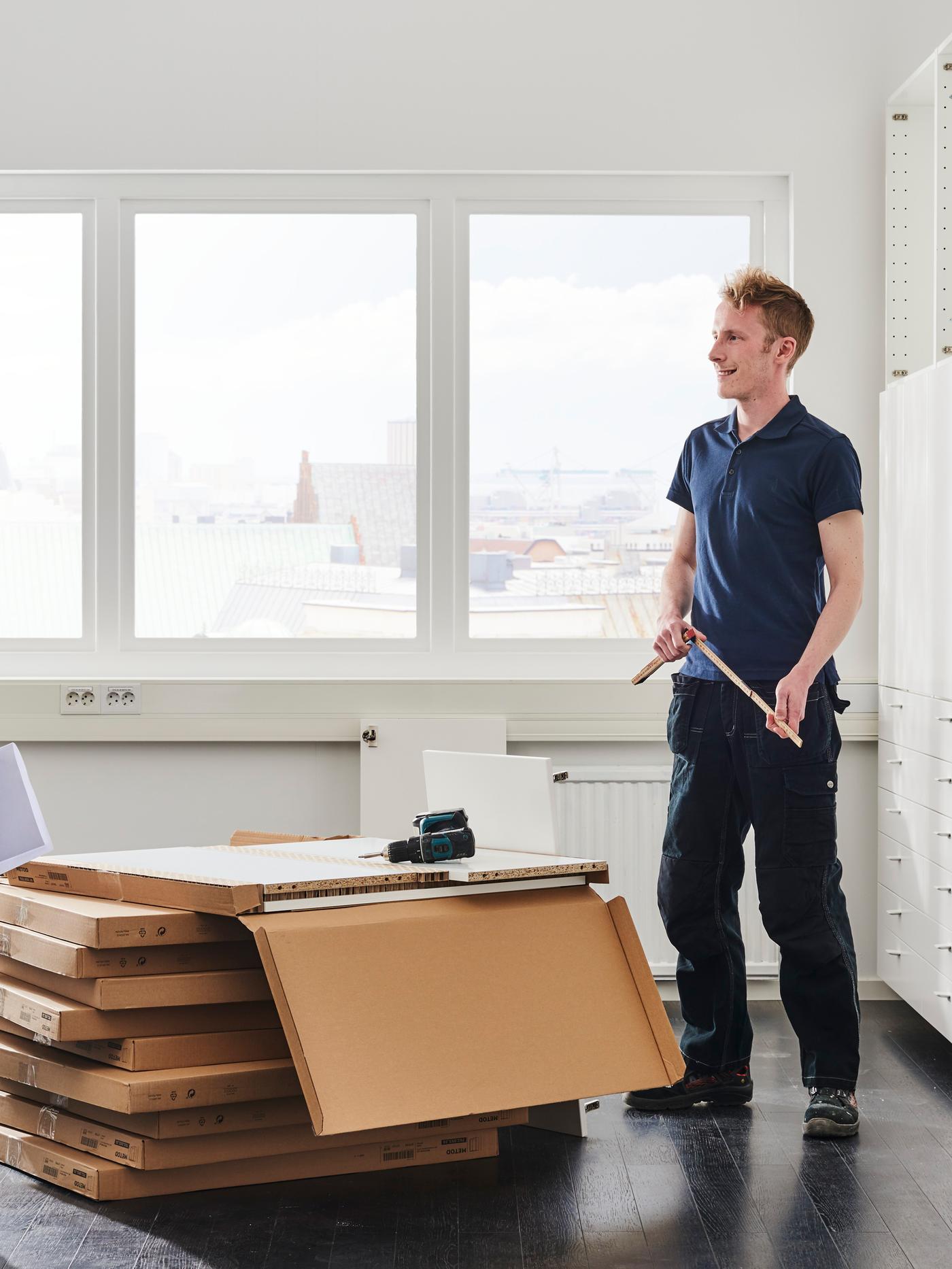A man stands by a pile of boxes with a tape measure in his hands, white walls and a window behind.