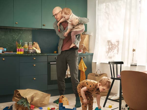 A man stands in a kitchen with dark green fronts and holds a baby in his arms. He looks startled because the shopping bag has fallen over. A toddler picks things up from the floor.