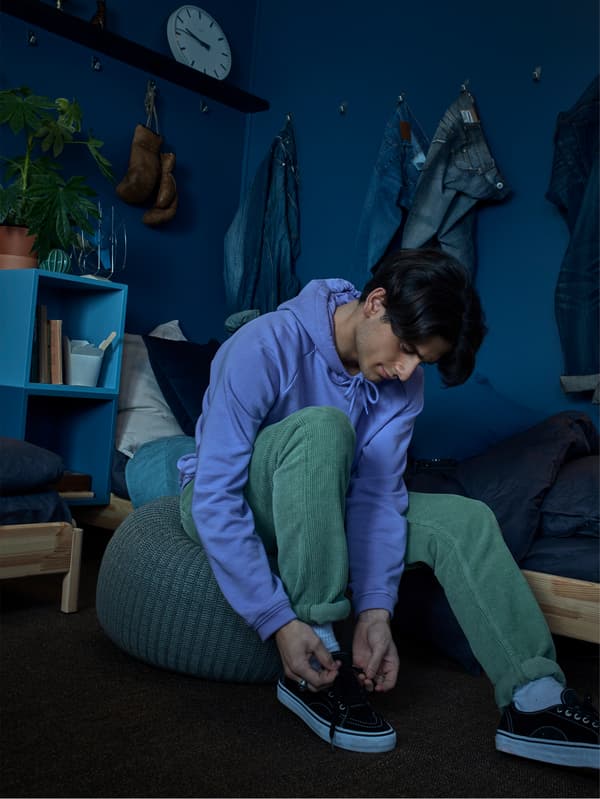 A man tying his shoes on a soft knitted SANDARED grey pouffe in a dark blue room.