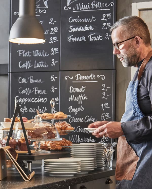 A man wearing a blue apron standing behind SÄLJAN worktop in black, with plates and jars, shown against a hand-written menu.