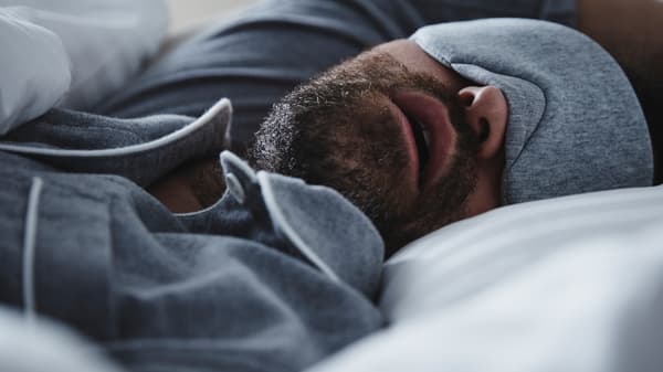 A man wearing grey pyjamas and a matching eye-mask is sleeping with his mouth open in a bed with NATTJASMIN bed linen.