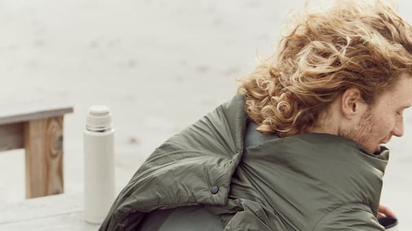 A man wears the FÄLTMAL quilt on the beach, secured at the back with a smart button fastening.
