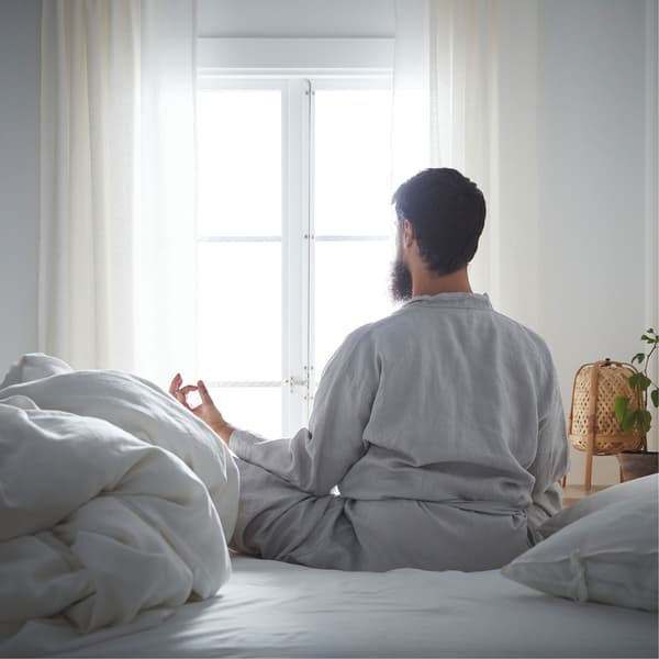 A man with a beard in a quiet room sitting with his legs crossed in a meditation pose with his back to the camera.