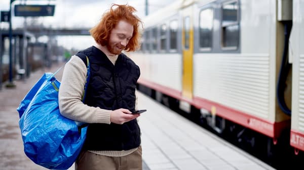 A man with a blue IKEA bag over his shoulder is standing on a train platform and looking at his smartphone.